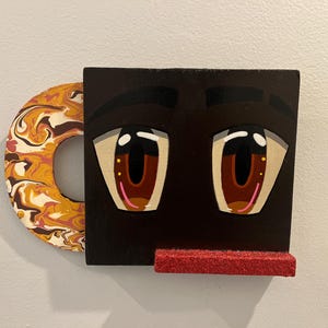 May include: A quirky wall decoration depicting a face with large, cartoon-like eyes. The eyes are brown with pink details, set against a black backdrop. A red, glittery shelf is below the eyes, and a circular handle with a marbled design is on the left.