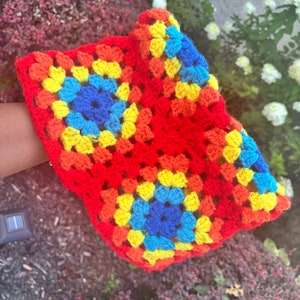 May include: A brightly coloured crochet hat, crafted in a granny square design. The hat showcases a striking palette, with red as the dominant hue, complemented by squares of yellow, orange, and blue. A unique and colourful accessory.