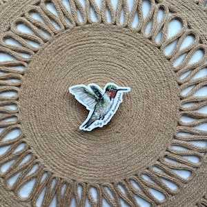 May include: A sticker of a ruby-throated hummingbird with green, red, and white details. The sticker is placed on a round, woven jute placemat with a decorative edge. The placemat is a natural tan color.