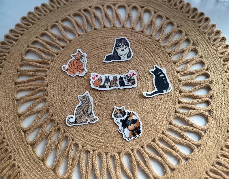 May include: A collection of cat stickers in various poses and colors, including orange tabby, black, and calico, arranged on a round, woven jute placemat. The stickers feature different cat breeds and designs, perfect for cat lovers.