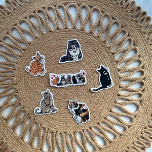 May include: A collection of cat stickers in various poses and colors, including orange tabby, black, and calico, arranged on a round, woven jute placemat. The stickers feature different cat breeds and designs.