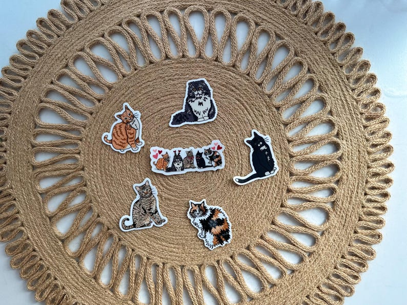 May include: A collection of cat stickers in various poses and colors, including orange tabby, black, and calico, arranged on a round, woven jute placemat. The stickers feature different cat breeds and designs.