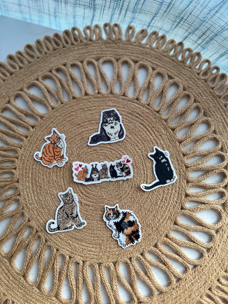 May include: A collection of cat stickers in various poses and colors, including orange, black, and calico. The stickers are arranged on a round, woven jute placemat. The cats are depicted in a cartoon style.
