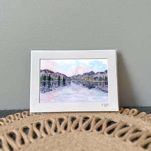 May include: A watercolor landscape painting of a serene lake scene, framed in a white mat. The artwork depicts mountains, trees, and a reflective body of water, with soft pink and blue hues. The artist's initials, K&N, are visible in the lower right corner.