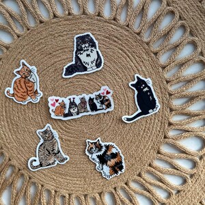 May include: A collection of cat stickers in various poses and colors, including orange tabby, black, and calico. The stickers are arranged on a woven, circular placemat with a textured surface.
