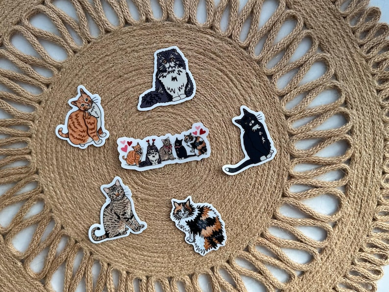 May include: A collection of cat stickers in various poses and colors, including orange tabby, black, and calico. The stickers are arranged on a woven, circular placemat with a textured surface.