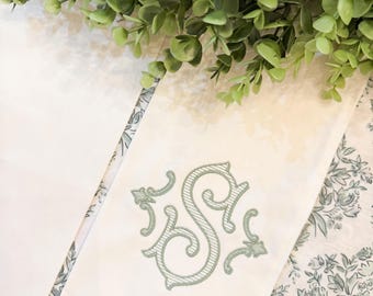 May include: Ivory-colored linen napkin with a decorative embroidered initial "S" in a soft green hue. The napkin is folded and placed on a floral patterned fabric, with a green leafy garland in the background. A classic and elegant design.