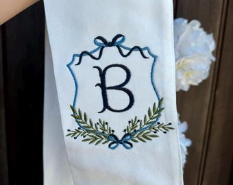 May include: White fabric with a blue and green embroidered monogram of the letter "B" in a shield shape with a bow at the top.