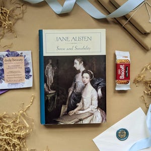 May include: A still-life arrangement featuring a copy of "Sense and Sensibility" by Jane Austen, a packet of tea, a Biscoff cookie, and a sealed envelope. The items are arranged on a tan surface with decorative paper and a light blue ribbon.