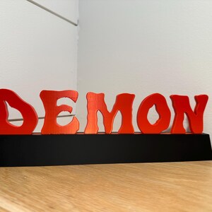 May include: A wooden sign that spells out the word "DEMON" in bright red, stylized letters. The letters sit on a black base, and the sign is displayed on a wooden surface.