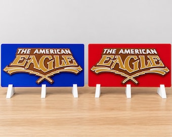 Puede incluir: Dos letreros rectangulares con el texto THE AMERICAN EAGLE en una fuente dorada superpuesta. Un letrero tiene un fondo azul sólido y el otro un fondo rojo sólido. Cada letrero se apoya sobre tres pequeños soportes triangulares blancos.