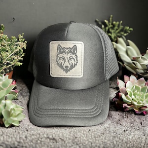 Puede incluir: Gorra de camionero gris carbón con parte trasera de malla y visera curva. La parte delantera presenta un parche cuadrado con un diseño detallado de cabeza de lobo. La gorra se muestra sobre una superficie gris texturizada, con plantas verdes en el fondo.