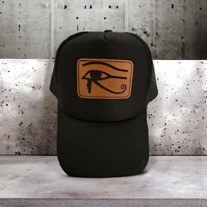 May include: Black trucker hat with a brown leather patch featuring a black Eye of Horus symbol. The hat has a mesh back and a curved brim. The patch is rectangular and centered on the front of the hat.