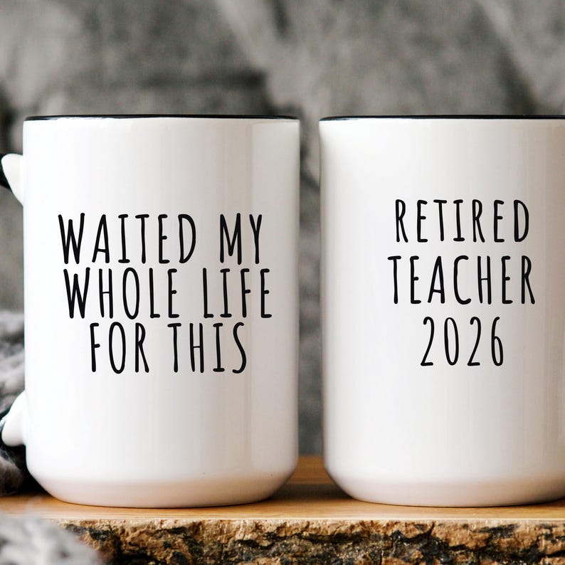 May include: Two white ceramic mugs with black rims. One mug reads "WAITED MY WHOLE LIFE FOR THIS" in black text. The other mug says "RETIRED TEACHER 2026". The mugs are on a wooden surface.