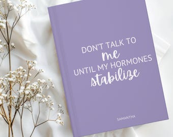 May include: A lavender journal with the text "DON'T TALK TO me UNTIL MY HORMONES stabilize" in white. The name "SAMANTHA" is printed at the bottom. The journal is on a white surface with white flowers.