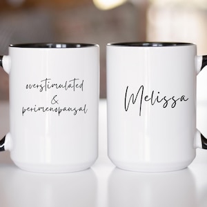 May include: Two white ceramic mugs with black interiors and handles. The left mug features the black text "overstimulated & perimenopausal" in a script font. The right mug features the black script name "Melissa" printed on the side.