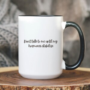 May include: White ceramic mug with a black handle and rim. The mug features the text "Don't talk to me until my hormones stabilize" in a black, handwritten-style font. The mug is set on a wooden surface.