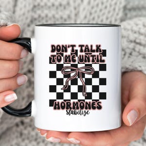 May include: White ceramic mug with a black handle and rim. The mug features the text "DON'T TALK TO ME UNTIL HORMONES stabilize" in black and pink lettering, with a pink bow graphic over a black and white checkered background.