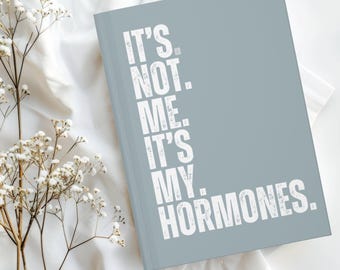 May include: A light blue journal with the text "IT'S. NOT. ME. IT'S MY. HORMONES." in white. The journal is closed and resting on a white surface, with delicate white flowers in the foreground.