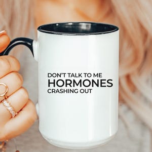 May include: White ceramic mug with a black interior and handle. The mug features the text "DON'T TALK TO ME HORMONES CRASHING OUT" in black, bold font. The mug is held by a person.