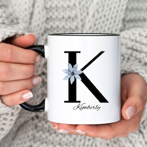 May include: White ceramic mug with a black rim and handle. The mug features a large black letter "K" with a blue flower on the left side of the letter. The name "Kimberly" is printed below the letter.