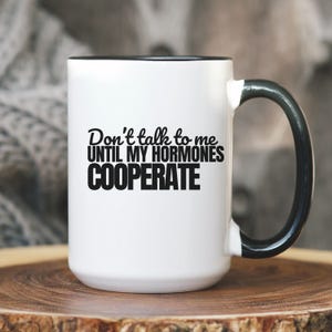 May include: White ceramic mug with a black interior and handle. The mug features the text "Don't talk to me UNTIL MY HORMONES COOPERATE" in black, bold lettering. The mug is sitting on a wooden surface.