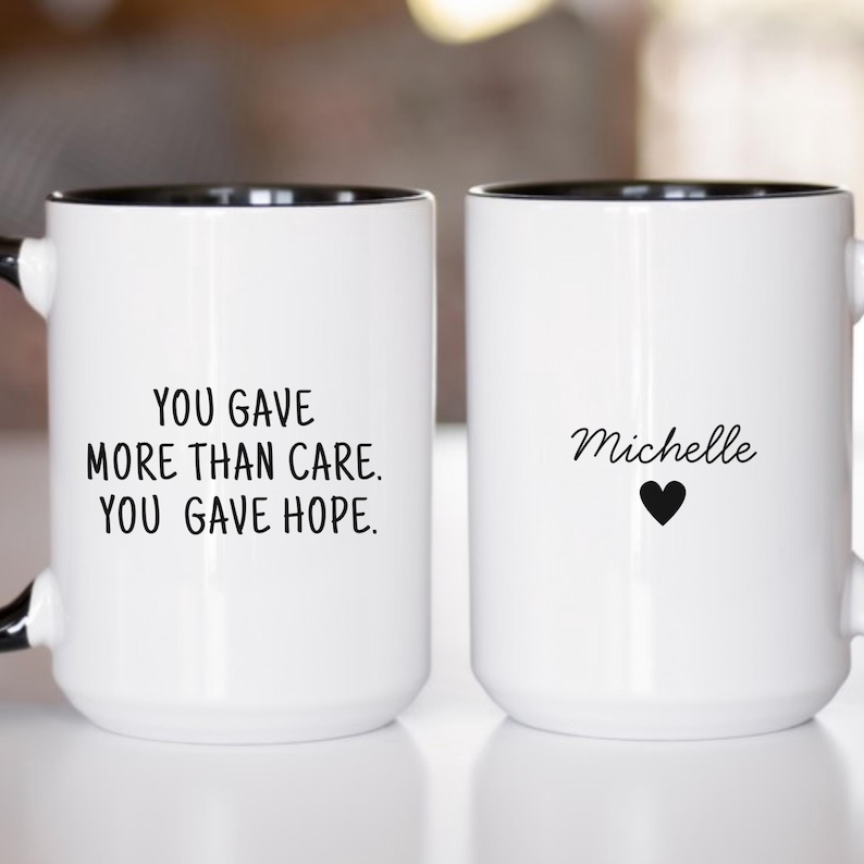Two white ceramic mugs with black interiors. Left mug reads: YOU GAVE MORE THAN CARE. YOU GAVE HOPE. Right mug displays the name Michelle above a small heart icon.