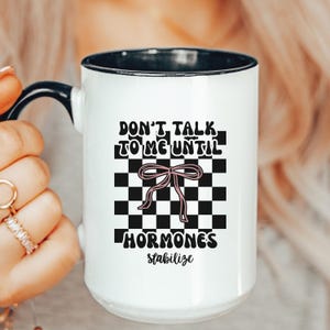 May include: White ceramic mug with a black rim and handle. The mug features the text "DON'T TALK TO ME UNTIL HORMONES stabilize" in a black, retro font, with a pink bow graphic on a black and white checkered background.