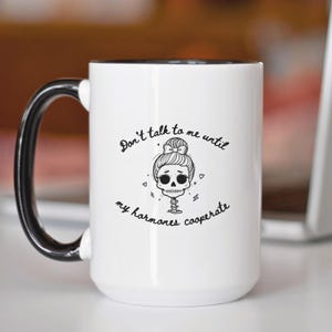 May include: White ceramic mug with a black handle and rim. The mug features a black and white illustration of a skull with a bun and the text "Don't talk to me until my hormones cooperate."