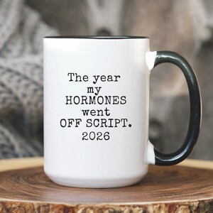 May include: White ceramic mug with a black interior and handle. The mug features the text "The year my HORMONES went OFF SCRIPT. 2026" in black lettering. The mug is sitting on a wooden surface.