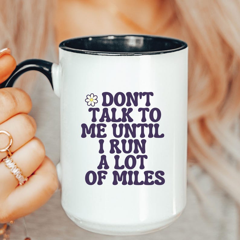 May include: White ceramic mug with a black interior and handle. The mug features the text "DON'T TALK TO ME UNTIL I RUN A LOT OF MILES" in purple, with a small daisy graphic. The mug is held by a person.