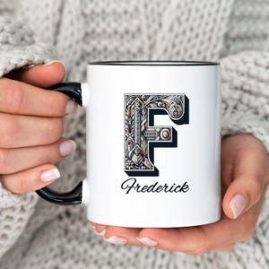 May include: White ceramic mug with a black rim. The mug features a stylized black and white illustration of the letter "F" with intricate details. The name "Frederick" is printed below the letter.