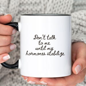 May include: White ceramic mug with a black interior and handle. The mug features the text "Don't talk to me until my hormones stabilize" in black script.
