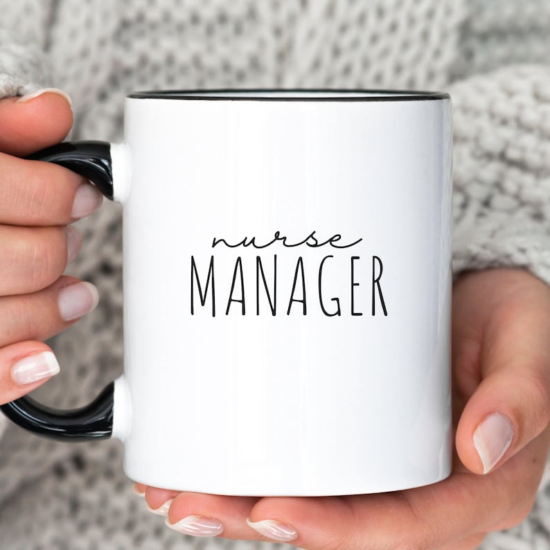 May include: White ceramic mug with a black handle and rim. The mug features the words "nurse MANAGER" in black lettering. The mug is held by a person wearing a gray sweater.