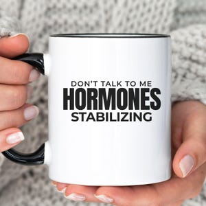 May include: White ceramic mug with a black interior and handle. The mug features the text "DON'T TALK TO ME HORMONES STABILIZING" in bold black letters. The mug is held by a person.