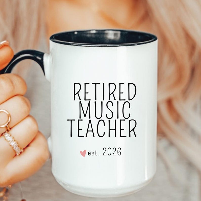 May include: A white ceramic mug with a black handle and interior rim. The front features black text that reads RETIRED MUSIC TEACHER with a small pink heart icon and the text est. 2026 printed in a simple sans-serif font on the center surface.