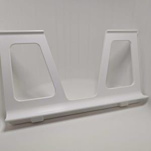 May include: A white plastic book stand featuring two trapezoidal side supports connected by a horizontal base ledge. The structure has a smooth finish with two large rectangular cutouts in the upright sections to hold items in an upright position.
