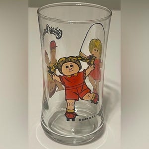 May include: Clear glass featuring a Cabbage Patch Kids design. The illustration shows a girl in a red outfit jumping rope, with other figures in the background. The glass has the text "Cabbage Patch Kids" and the copyright year "© 1984 O.A.A."