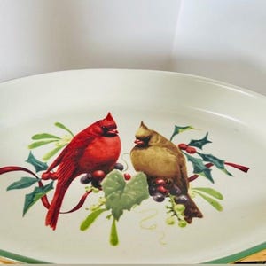 May include: Oval serving platter with a cream-colored surface and a green rim. The center features a detailed illustration of two cardinal birds, one red and one brown, surrounded by holly and berries.