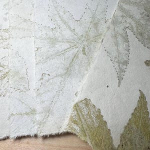 May include: Close-up of handmade paper with leaf imprints. The paper is off-white with visible texture and frayed edges. The leaf impressions are in shades of green and brown, creating a natural, botanical design.