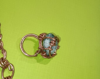 May include: A handmade copper ring featuring a wire-wrapped, oval-shaped, green and white stone. The ring has a simple band and a decorative wire cage around the stone. A copper chain is partially visible in the image.