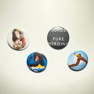 Könnte beinhalten: Vier runde Knöpfe mit verschiedenen Bildern. Der erste Knopf zeigt ein Schwarzweißbild einer Frau mit einer Katze. Der zweite Knopf hat einen schwarzen Hintergrund mit weißem Text, der "LORDE PURE HEROINE" lautet. Der dritte Knopf hat einen blauen Hintergrund mit einer Frau in einem gelben Kleid. Der vierte Knopf hat einen blauen Hintergrund mit einer Person, die in die Luft springt.