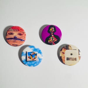 May include: Four round buttons with unique designs. One features a surreal landscape with eyes, another a purple silhouette filled with colorful shapes, a third a Rubik's Cube against a blue sky, and the last a person's face with text.