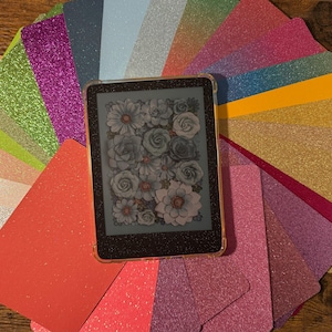 May include: A variety of glitter cardstock in different colors, including green, pink, purple, blue, gold, and red. The cardstock is arranged in a fan shape around a black e-reader with a floral design on the screen.