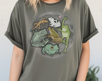 May include: A green t-shirt with a circle design featuring five frogs and a lizard. The frogs are green, brown, and white. The lizard is green and brown. The circle is on a green background.