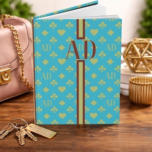 May include: A teal journal with a playing card pattern of hearts, diamonds, and clubs. The journal has the initials "AD" in a large font on the cover, with a brown vertical stripe. A pink purse and keys are in the background.