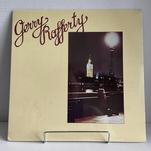 Peut inclure: Une pochette d'album de disque vinyle vintage avec une photo d'une personne debout sur un pont devant une ville nocturne. La pochette de l'album est jaune avec le texte "Gerry Rafferty" en lettres rouges.