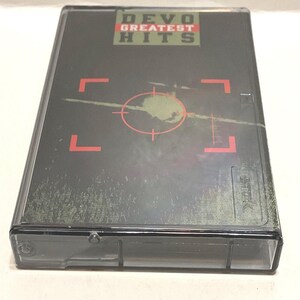 May include: A vintage cassette tape case with the text "DEVO GREATEST HITS" in red and green. The case has a dark gray background with a red target design and a faded, textured appearance.