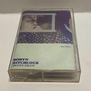 May include: A vintage cassette tape in a clear plastic case. The cassette label features a portrait and the text "ROBYN HITCHCOCK GROOVY DECOY" in blue. The background has a mix of colors, including purple, blue, and white.