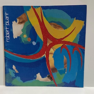 May include: Album cover for Robert Plant. The artwork is a collage of blue, yellow, and red shapes with a mirrored surface. The text "robert plant" is printed vertically in white on the left side. The design is abstract and colourful.